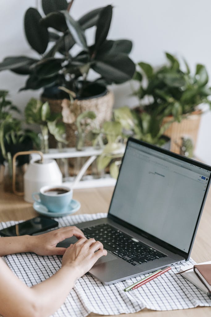 Crop anonymous freelancer browsing modern netbook while sitting at table in light room with many green potted plants at home